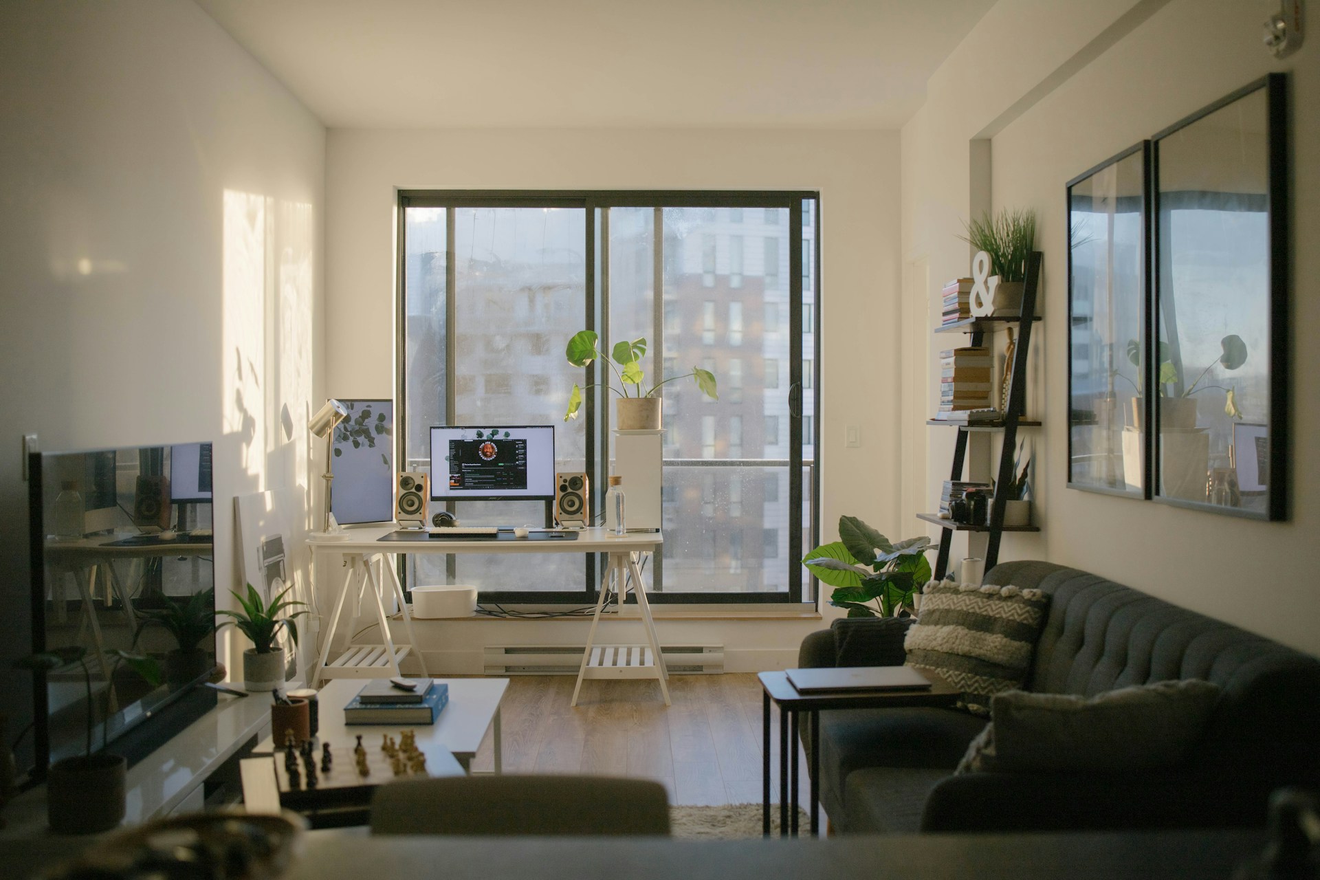 A modern home office with a dual-monitor desk, shelving, and a sofa, overlooking the city